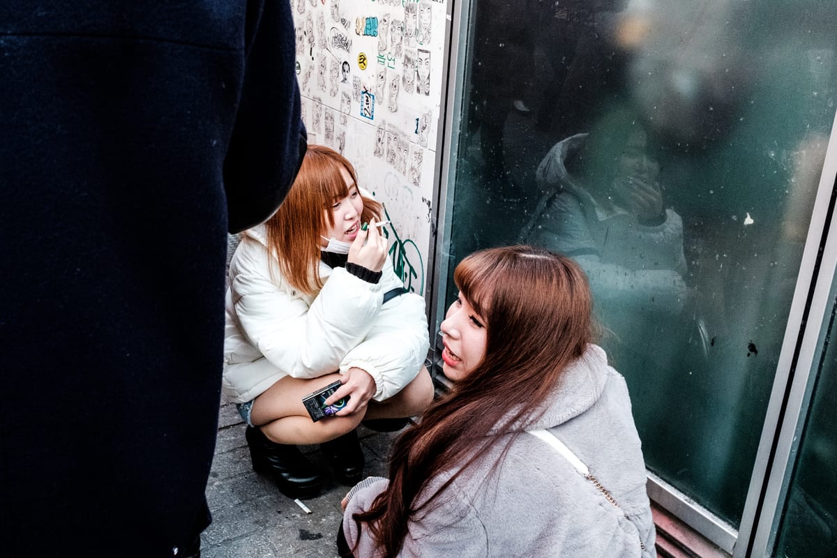 Two young women crouching against a glass storefront covered in stickers, one applying lipstick