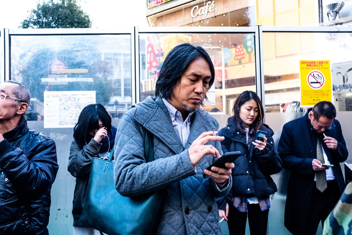 Man with long dark hair using his smartphone outside a cafe window, surrounded by others also on their phones