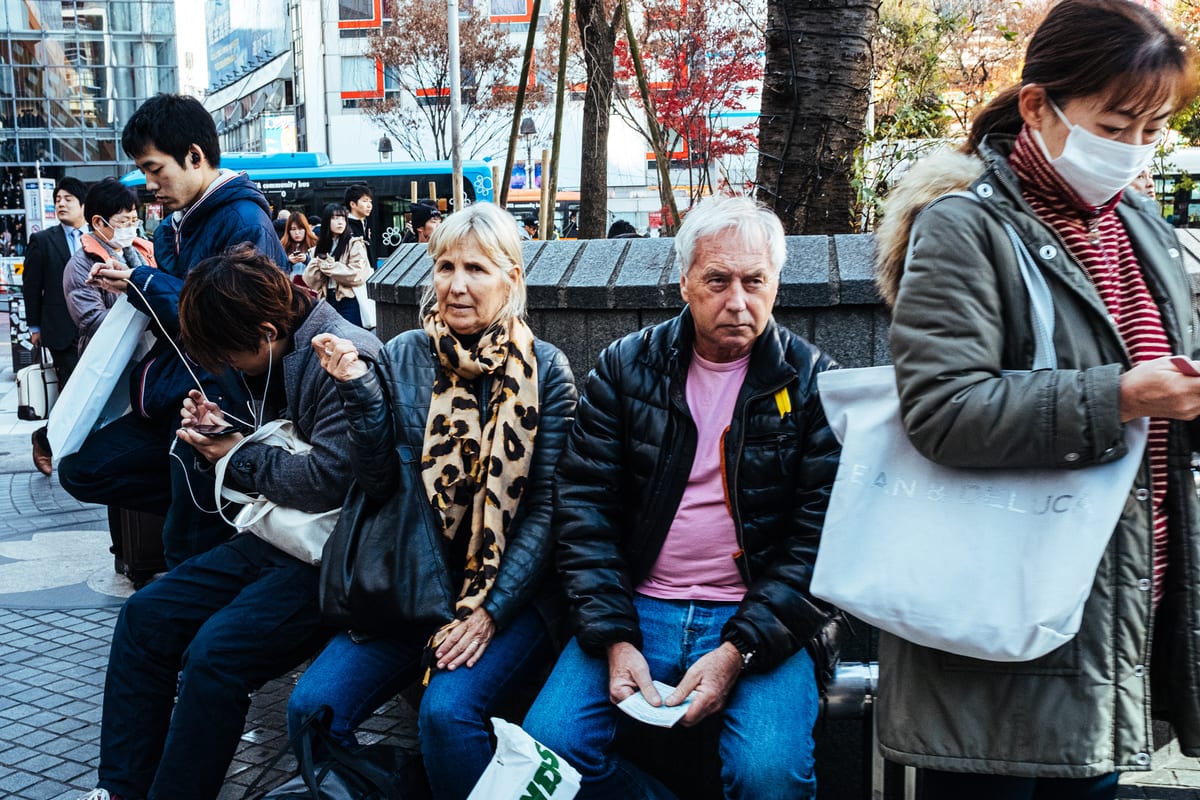 Older couple sitting on a bench among pedestrians, the woman in a leopard-print scarf and the man in a puffer jacket