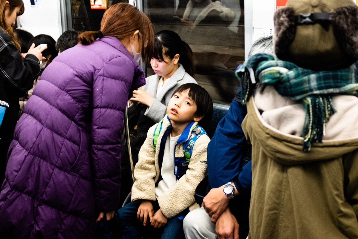 Young boy looking up from his seat on a crowded subway car as a woman in a purple jacket leans over him