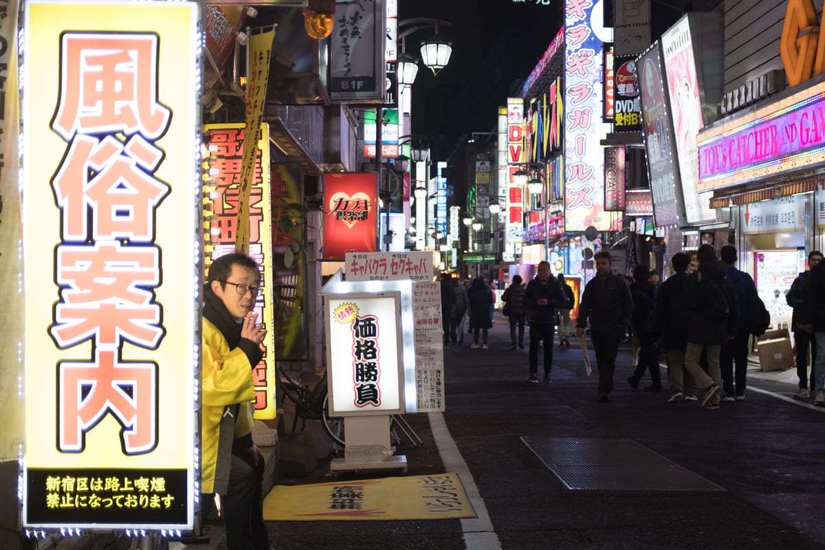Man sitting beside a large yellow sign with Japanese text on a neon-lit Kabukicho alley at night