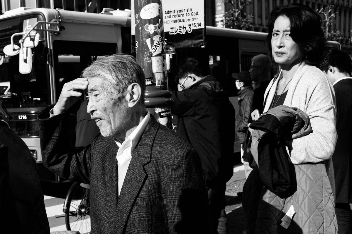 Elderly man in a tweed jacket scratching his head beside a woman in a kimono among pedestrians