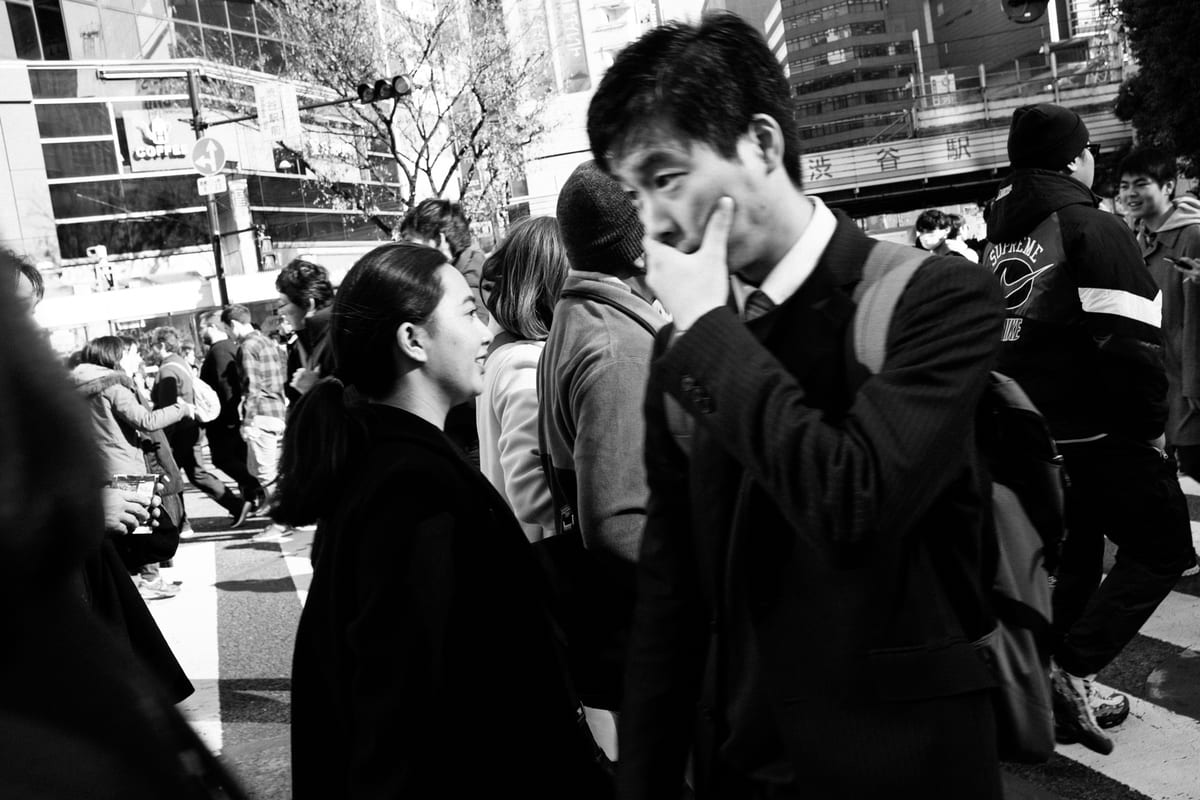 Man in a suit covering his face with his hand while a woman in a black coat looks toward him in a busy street crowd