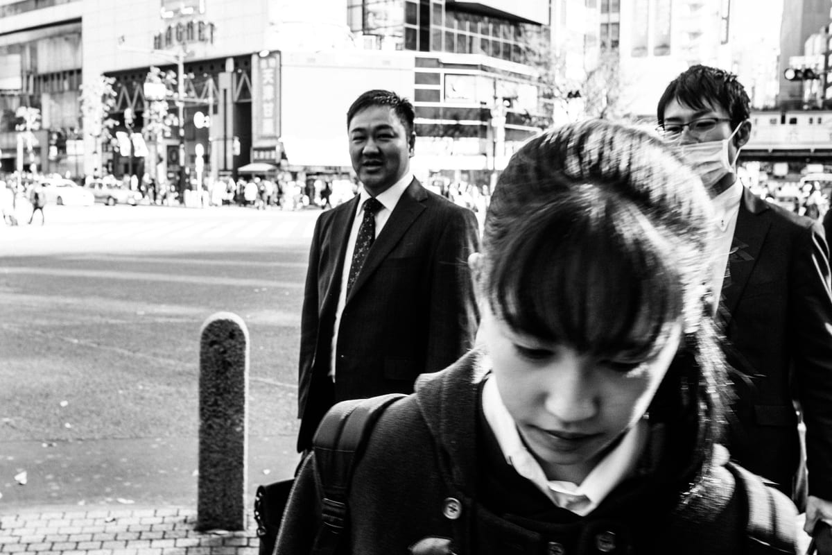 Young woman looking downward with two men in suits and face masks walking behind her on a city sidewalk