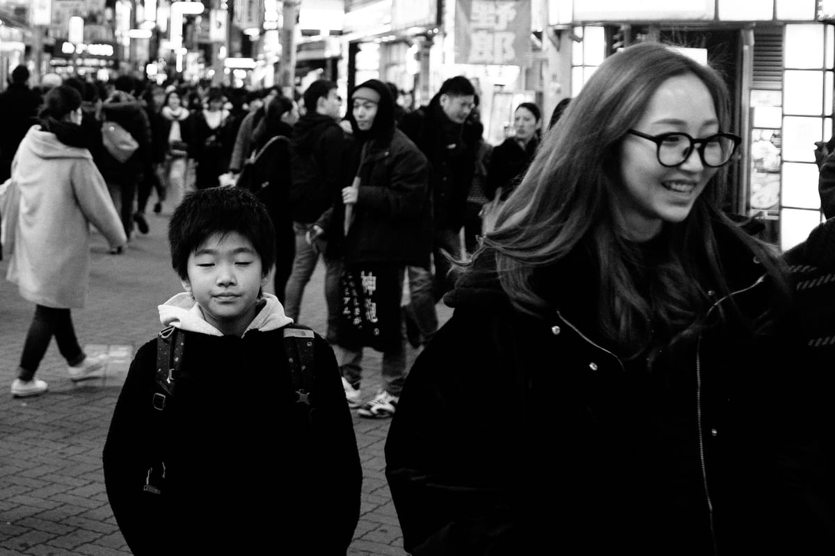 A boy and a smiling young woman in glasses walking on a crowded evening street with illuminated signs behind them