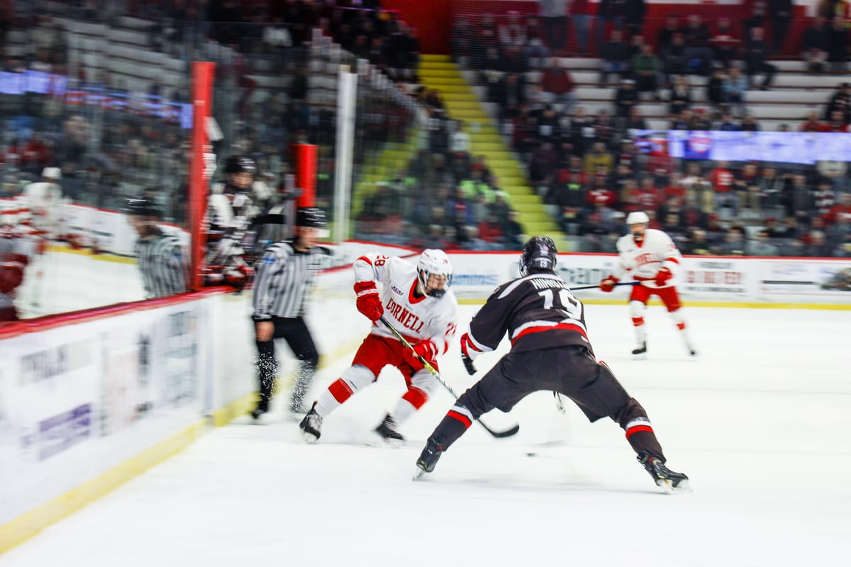 Cornell player in red battles for the puck against an opponent along the boards, referees visible in the background