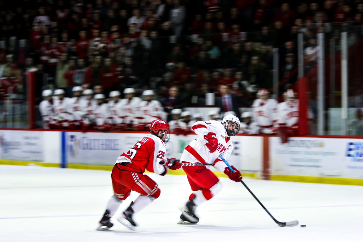Two Cornell players skate in tandem, one carrying the puck, with the team bench and crowd visible behind the glass