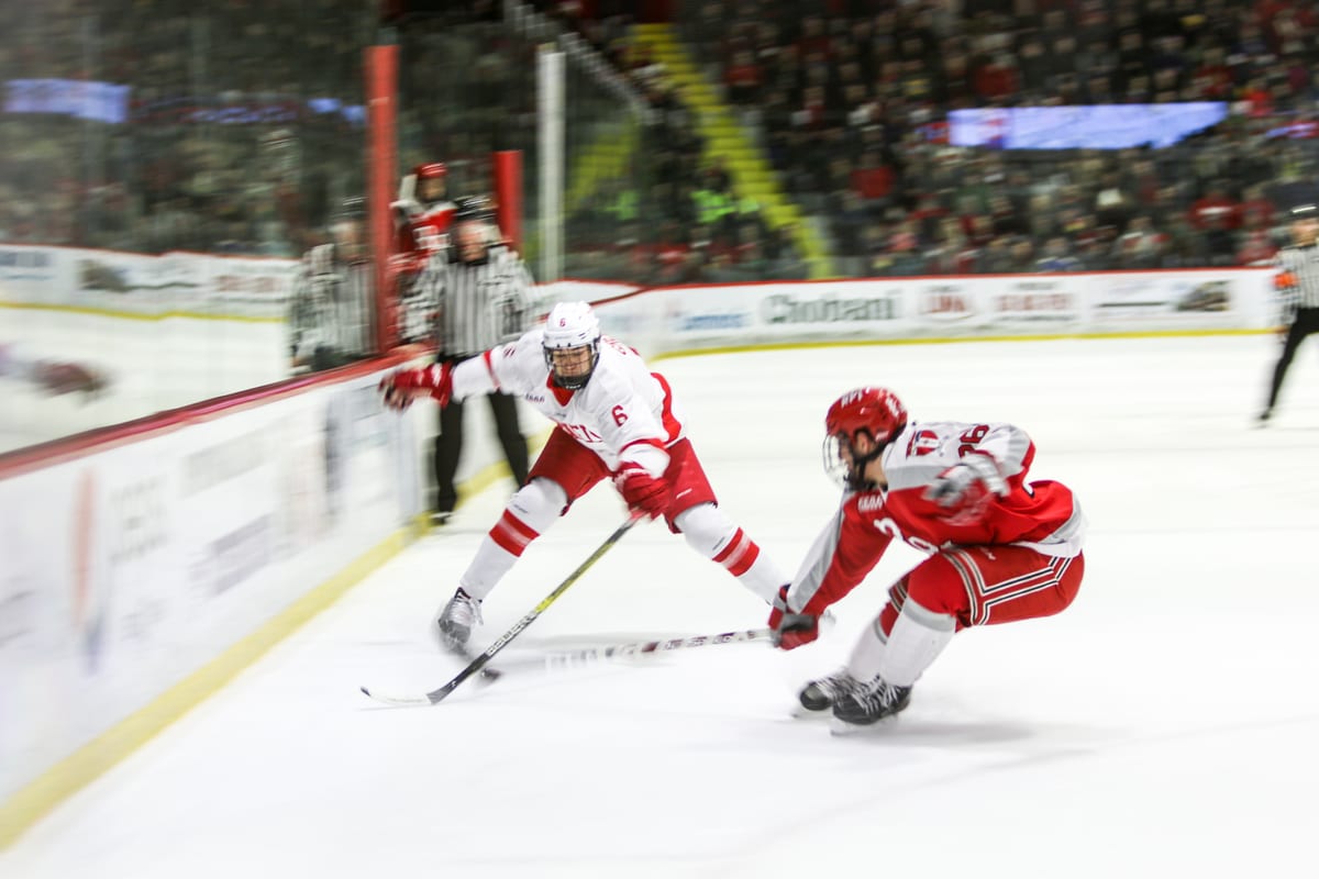 Cornell player extends to reach for the puck while an opponent lunges low to the ice, referee and crowd in the background