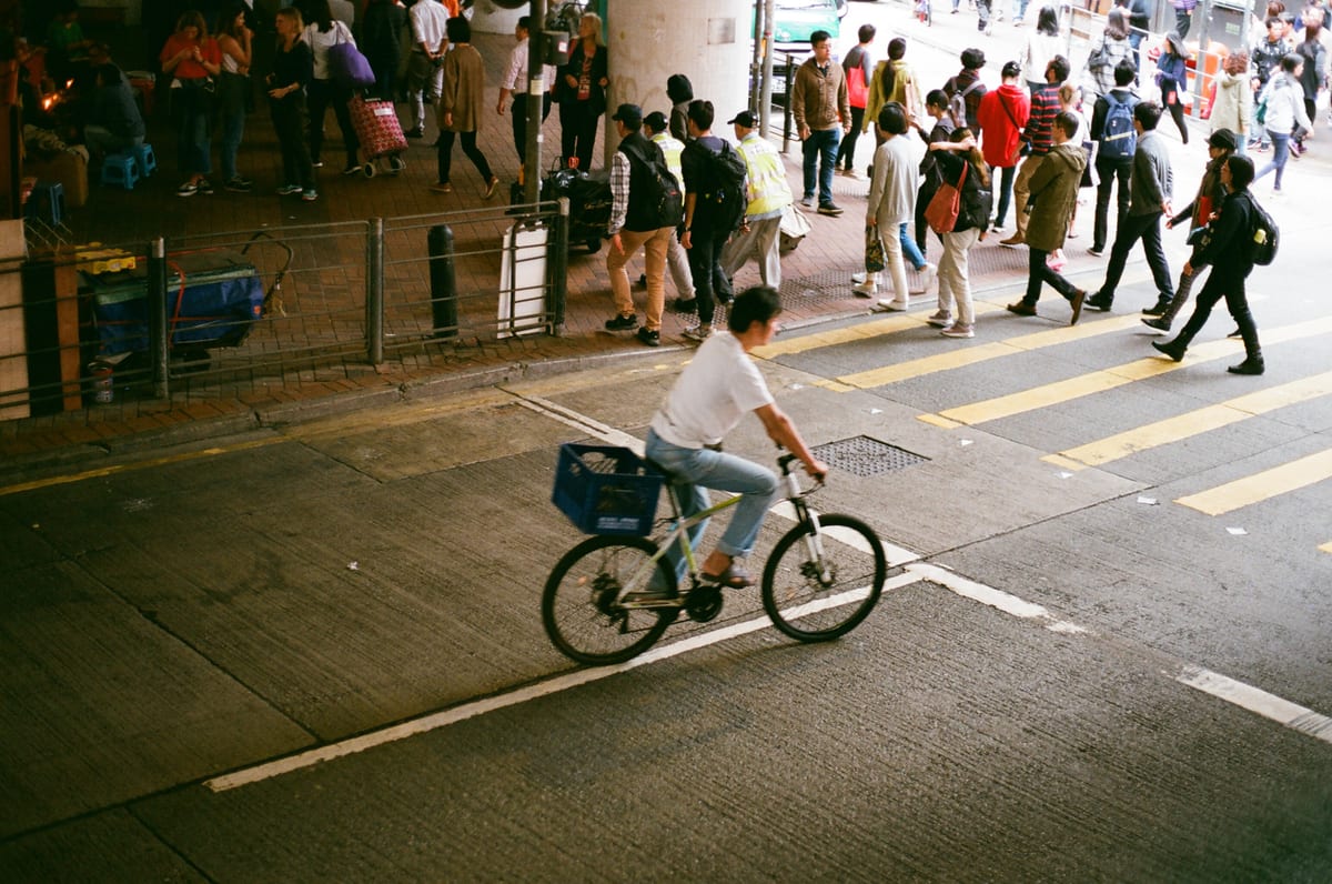 A cyclist with a front basket rides past a busy crosswalk where crowds of pedestrians cross under an overpass