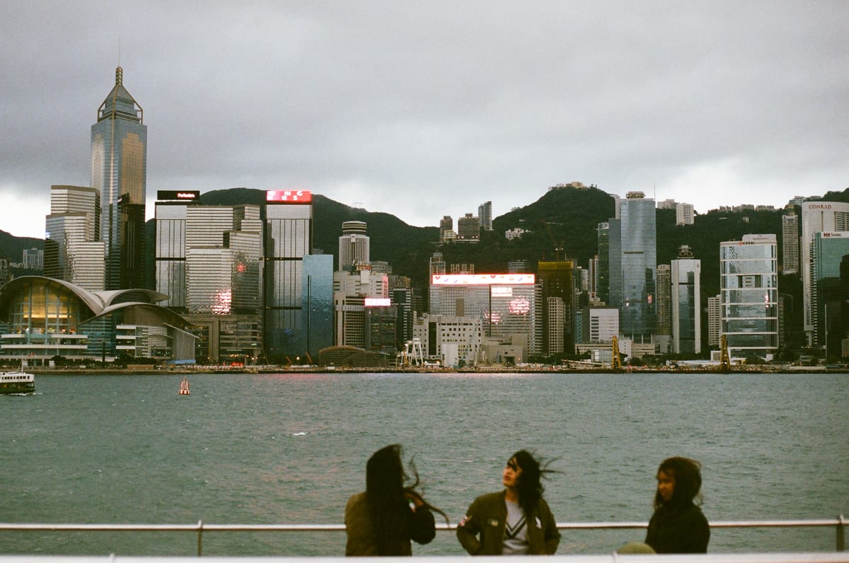 Three women standing at the waterfront with the Hong Kong Island skyline across the harbour, wind blowing their hair