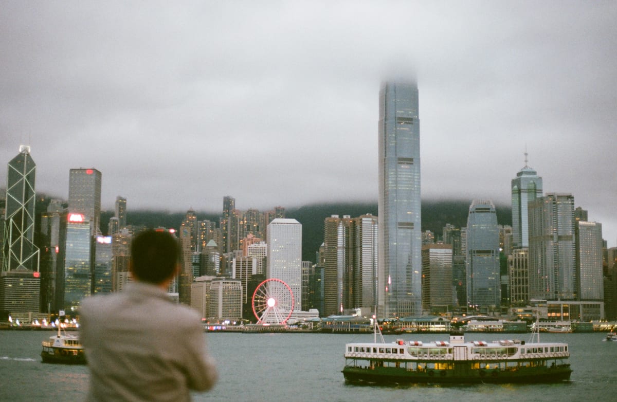 A man seen from behind looks across the harbour toward the Hong Kong skyline with ICC tower disappearing into low clouds, a Star Ferry crossing