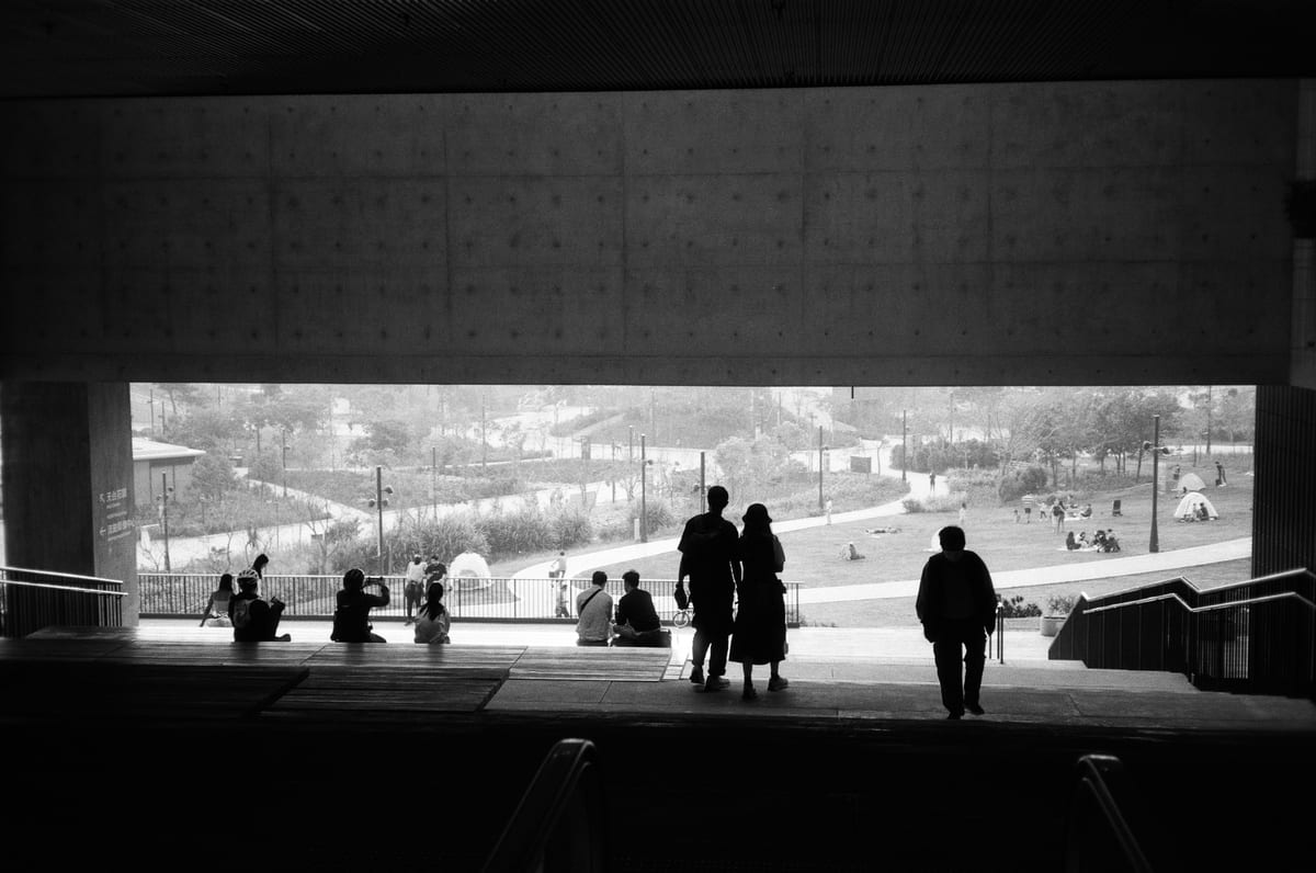 Silhouetted figures standing and sitting under a large concrete overhang, looking out toward a sunlit park with winding paths