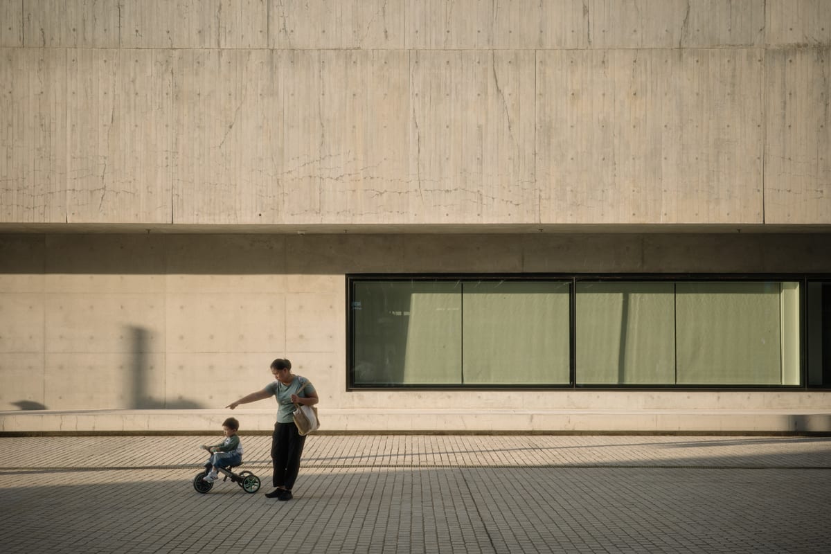 A man walks with a small child on a balance bike along a sidewalk in front of a large concrete building