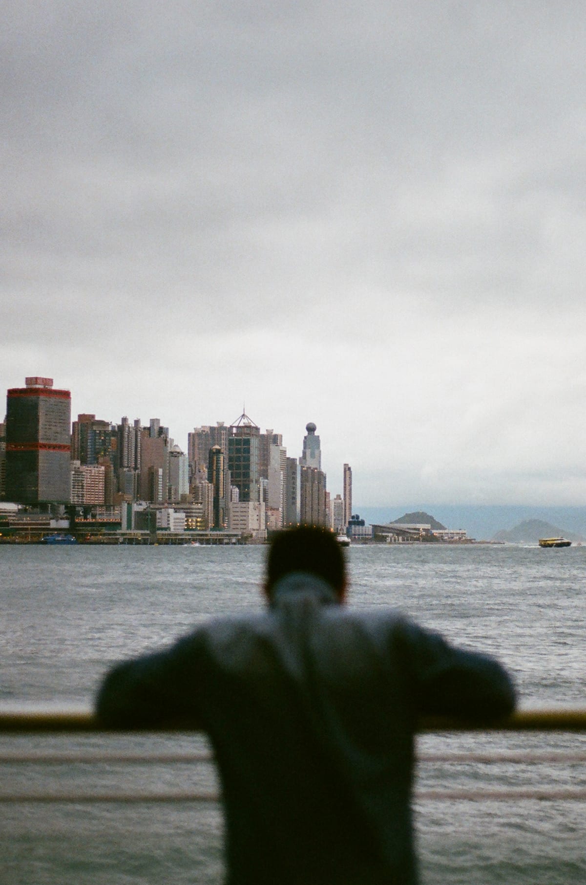 A man seen from behind leans on a railing, looking across the harbour toward the dense Hong Kong Island skyline under overcast skies