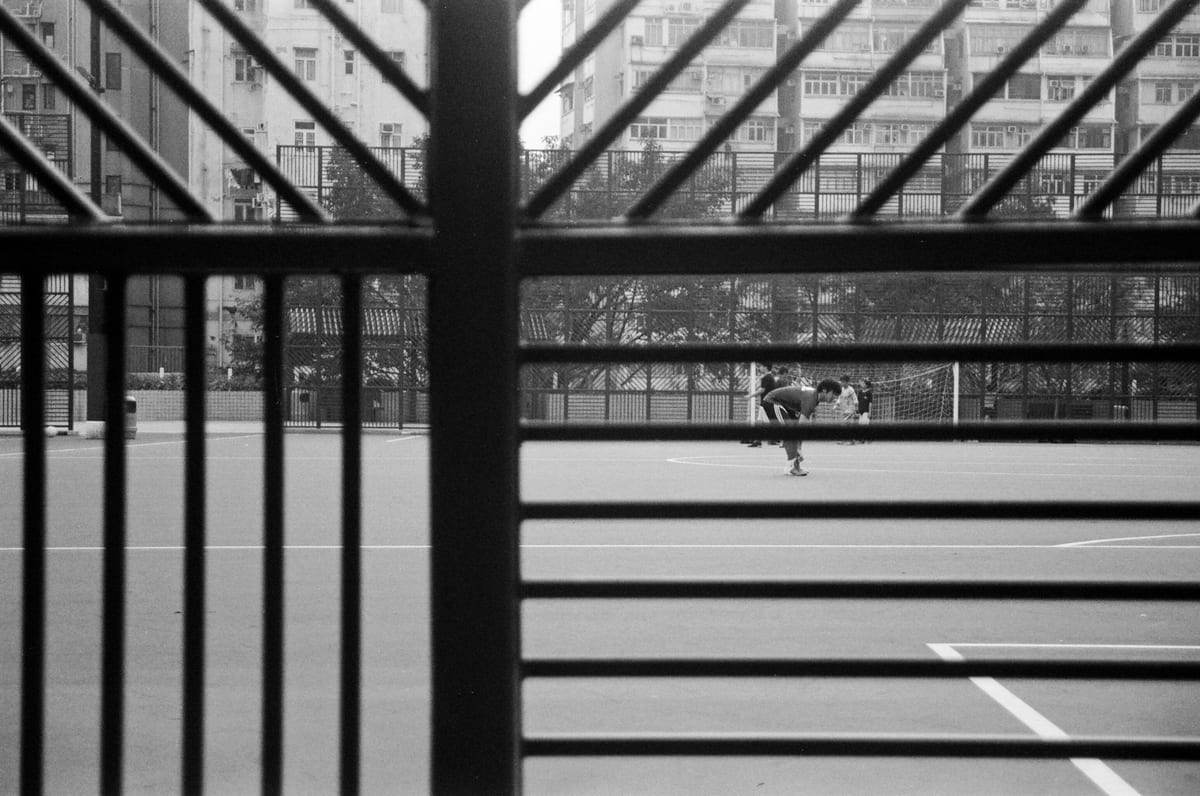 Two people playing on an outdoor sports court seen through metal railings with residential high-rises in the background