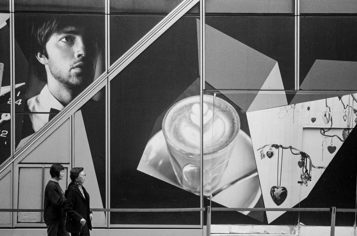 Two people walk past a large building-sized advertisement seen through diagonal escalator handrails