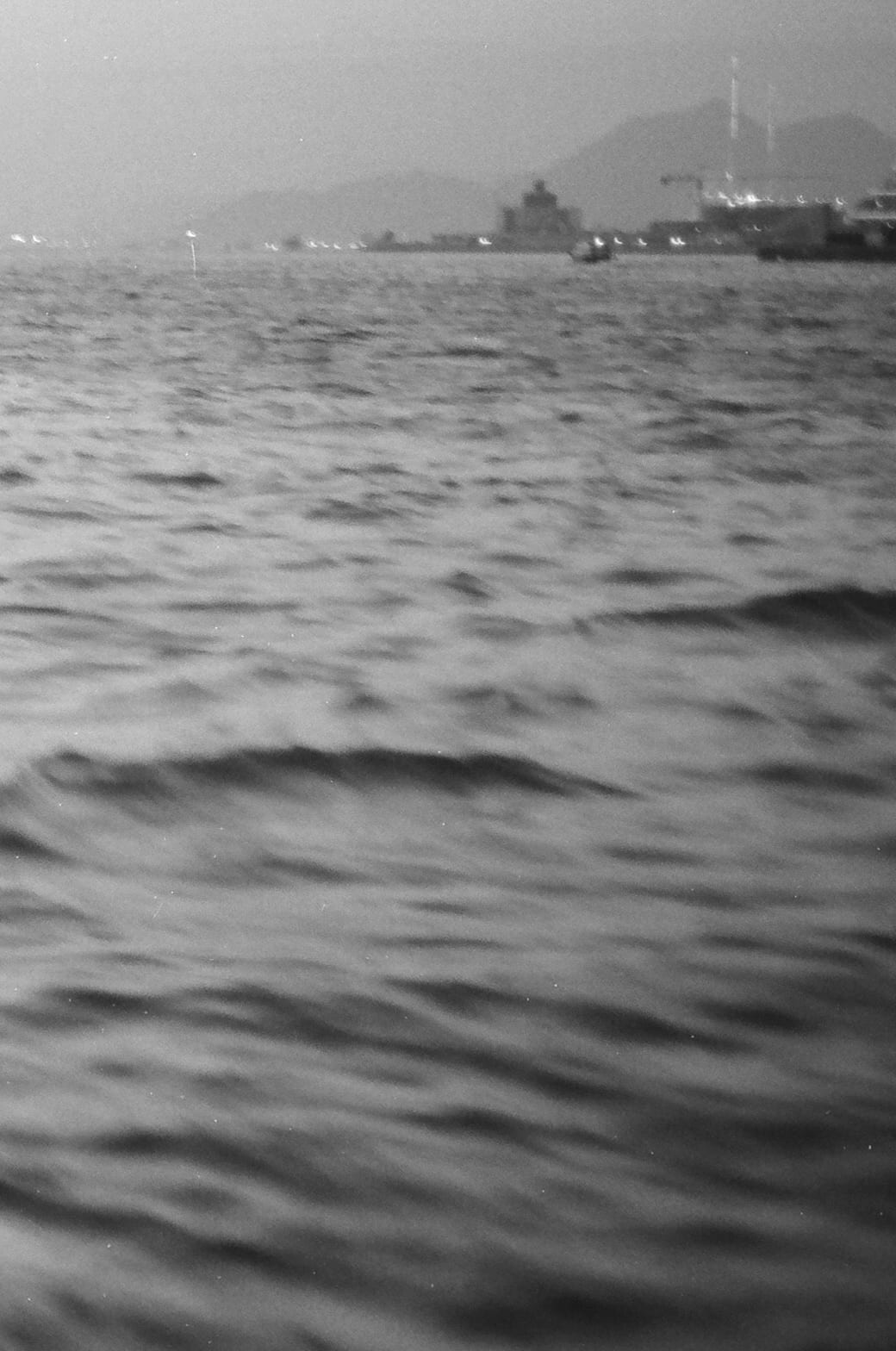 Choppy harbour water in the foreground with distant ships, hills, and hazy sky in the background
