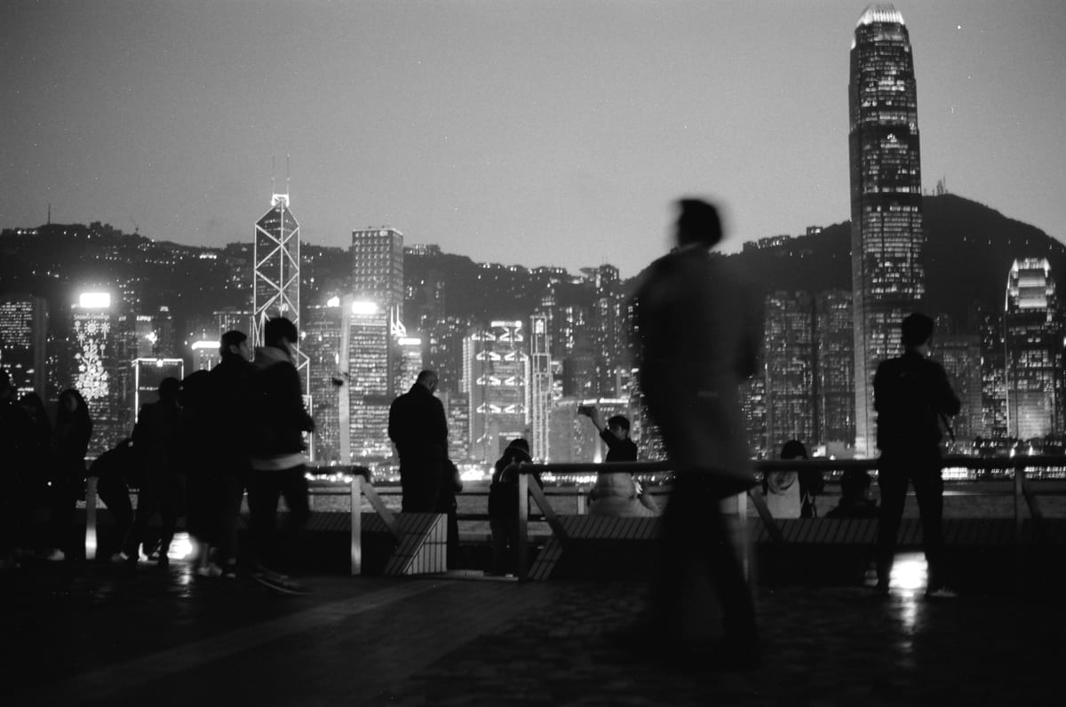 Silhouetted figures on a waterfront promenade at night with the illuminated Hong Kong Island skyline in the background