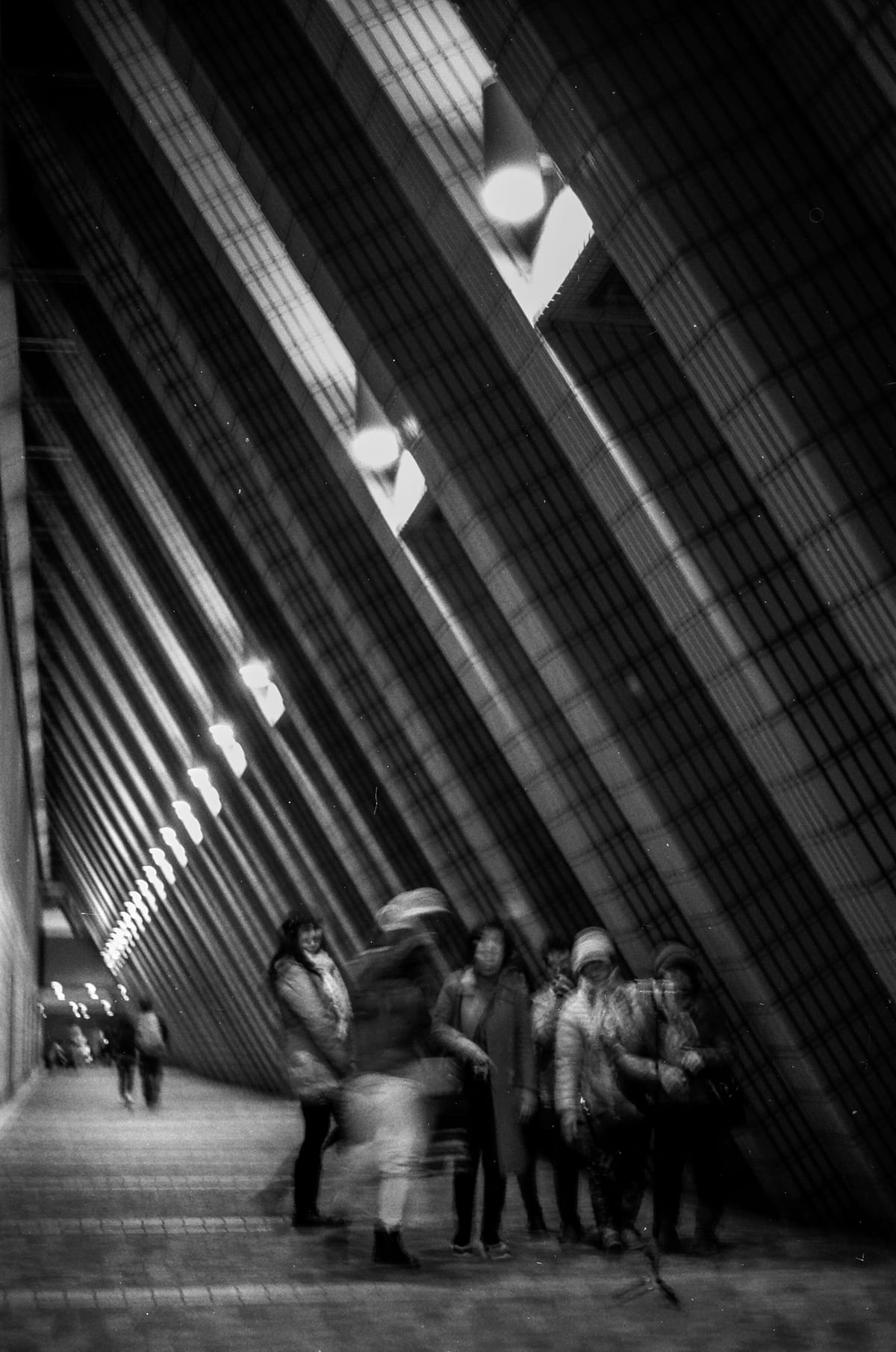 Motion-blurred pedestrians walking through a tall narrow corridor with dramatic converging lines of ribbed walls and ceiling lights