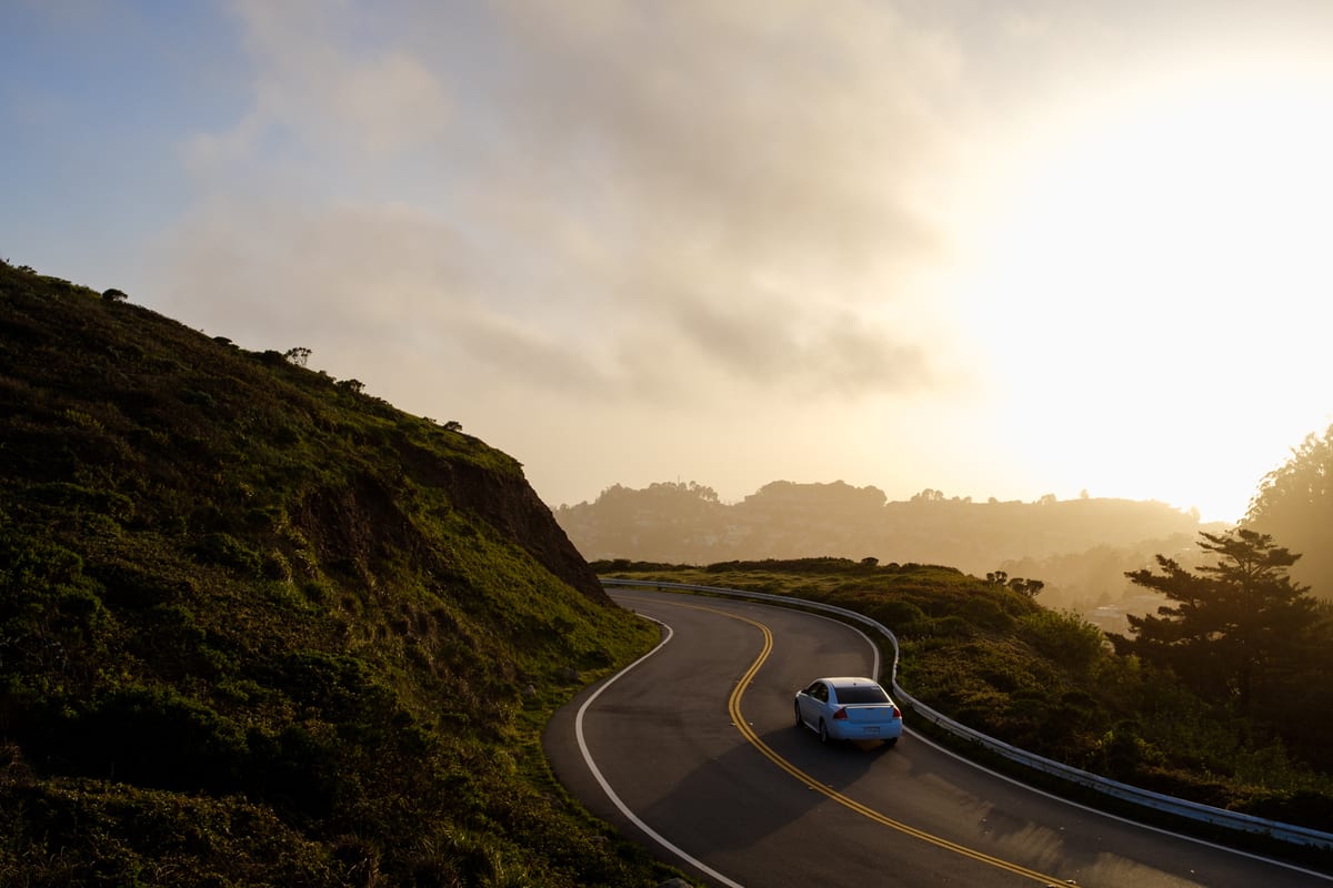 White car driving along a winding coastal road through green hills at sunset with fog in the distance