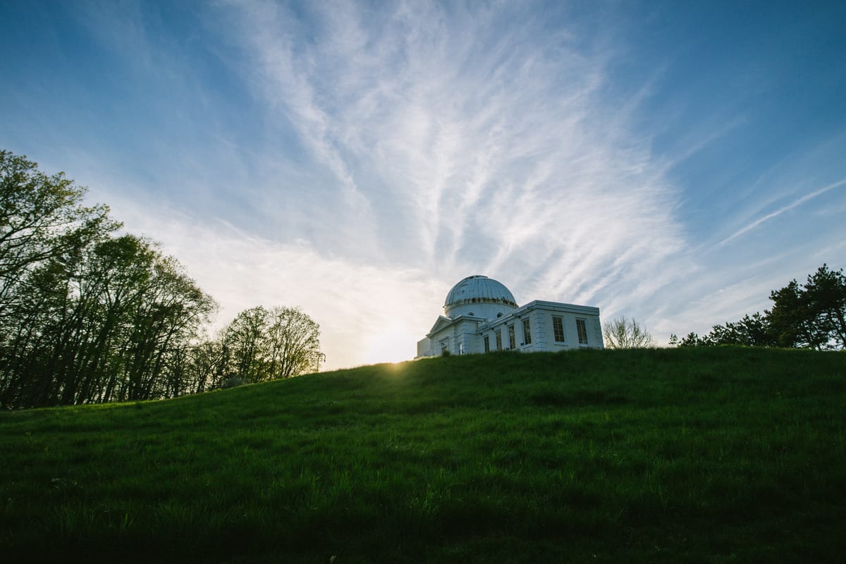 White domed observatory on a green hilltop silhouetted against a blue sky with wispy clouds at sunset