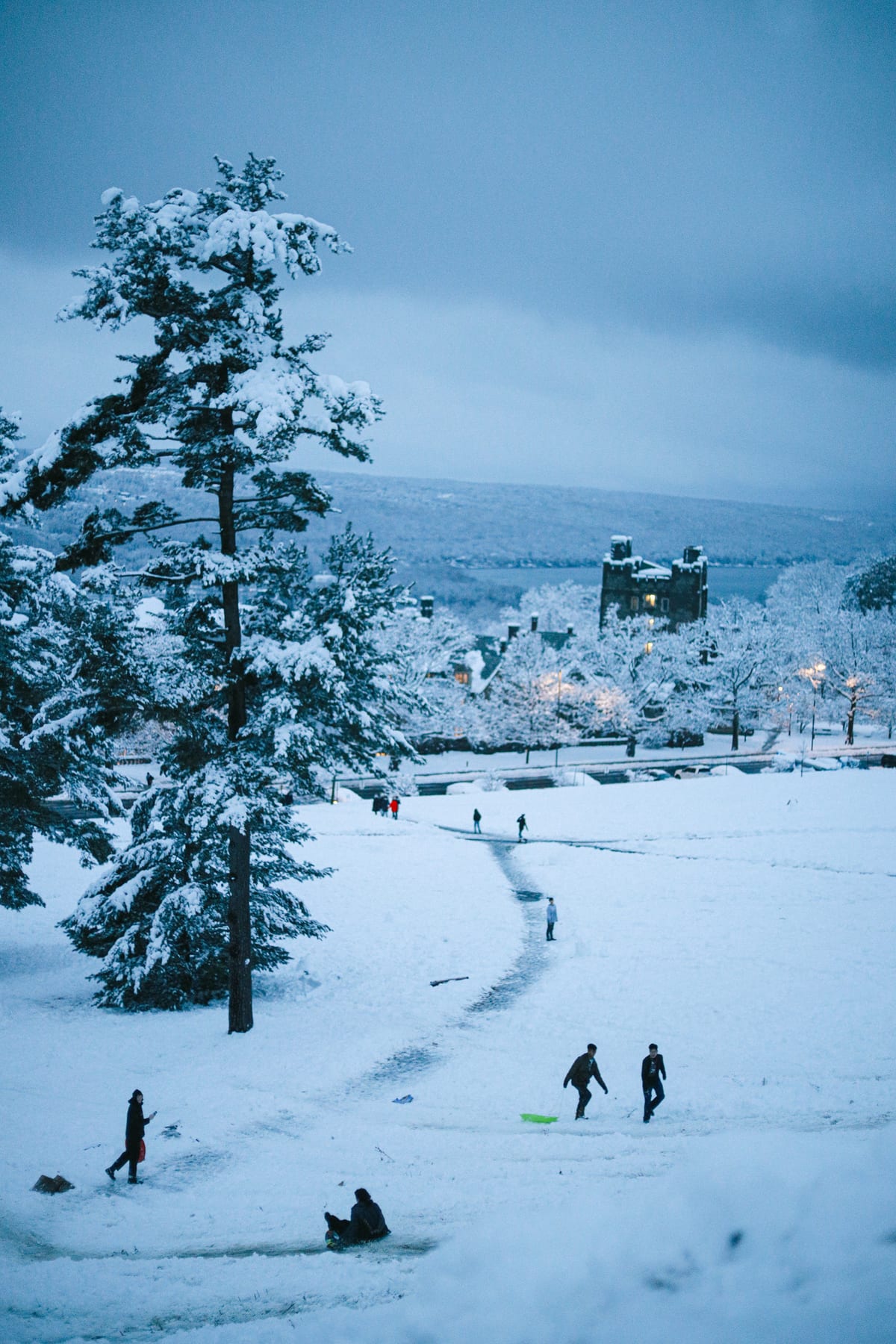 Snow-covered hillside at dusk with people walking and sledding, evergreen trees on the left and lit buildings in the valley below