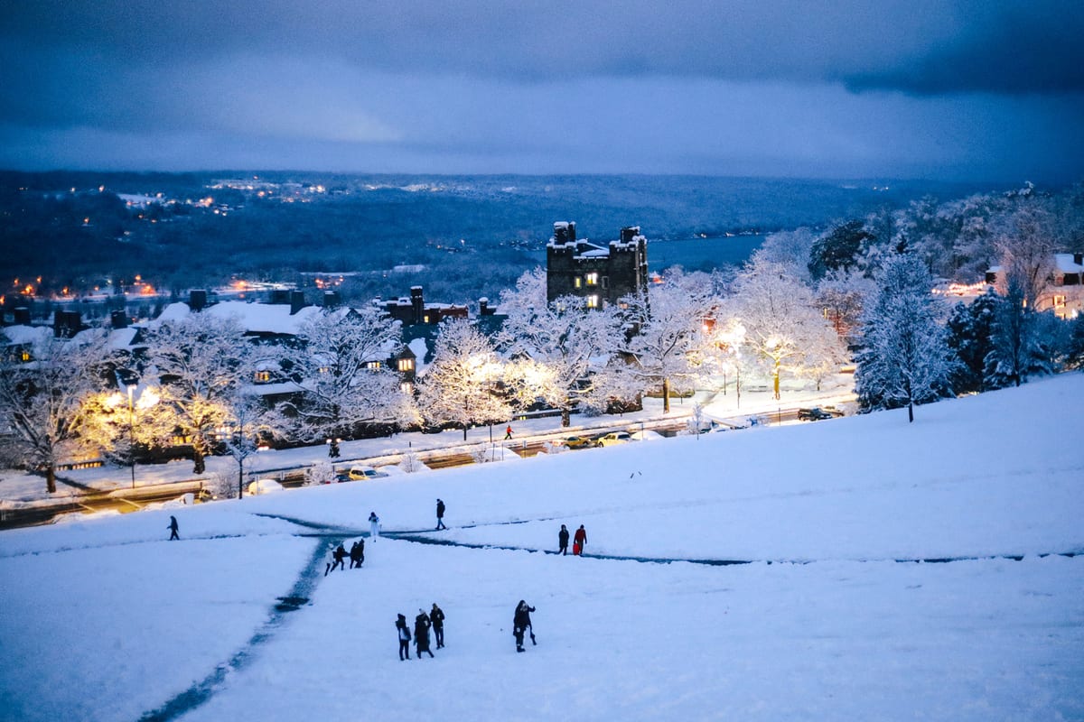 Snow-covered hillside at twilight with groups of people on paths, illuminated trees, and town lights in the valley